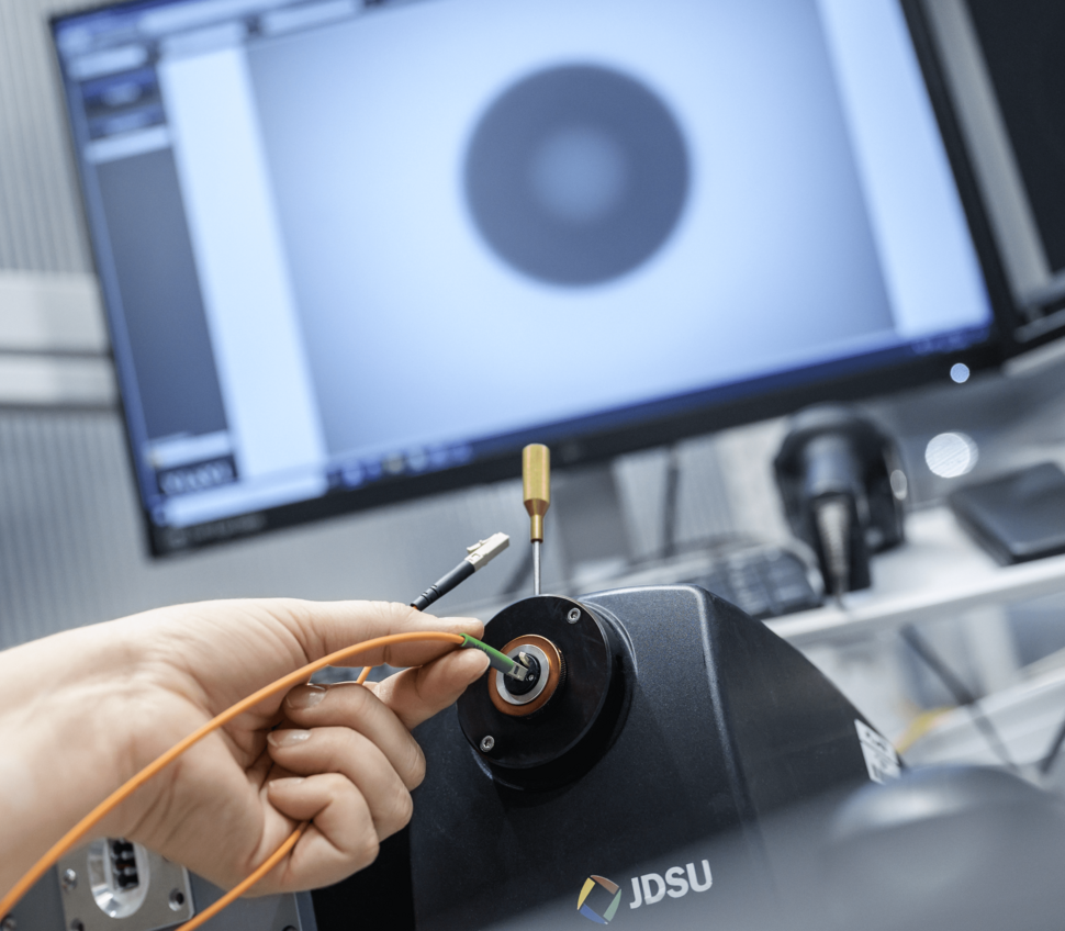 A hand holds a fiber optic cable end in a measurement device for surface testing.